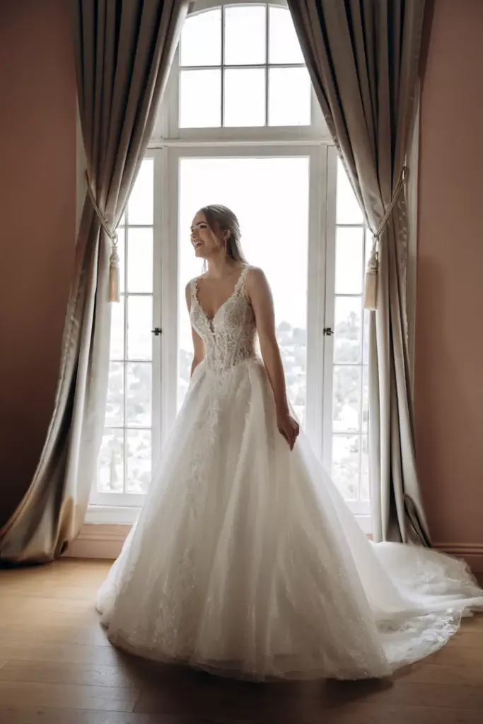 Brautkleid mit Spitzenoberteil, V-Ausschnitt und weiter Schleppe am Fenster
